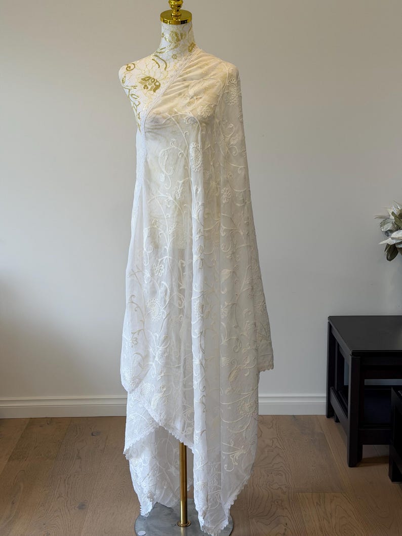 Pu&ograve; includere: Una lunga stola in tessuto bianco semi-trasparente caratterizzata da un intricato ricamo floreale tono su tono su tutta la superficie. Il materiale leggero cade con eleganza, mostrando delicati motivi botanici e un bordo smerlato.