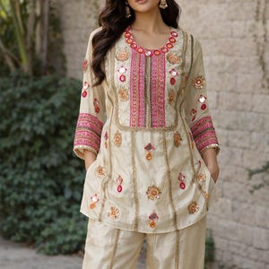 Peut inclure: Ensemble tunique et pantalon brodé de couleur crème. La tunique présente des broderies dorées et roses complexes autour de l'encolure, des manches et du corsage, avec de petits miroirs. Le pantalon est de coupe large.