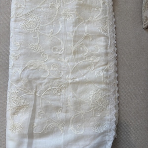 Pu&ograve; includere: Un pezzo di tessuto rettangolare color bianco sporco caratterizzato da un intricato ricamo floreale tonale su tutta la superficie. I bordi sono rifiniti con una delicata bordura in pizzo smerlato lungo i lati e il fondo dell'articolo.