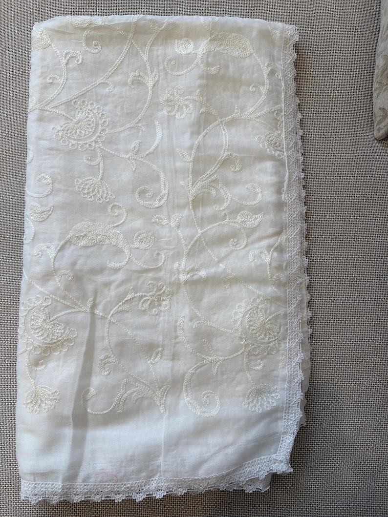 Pu&ograve; includere: Un pezzo di tessuto rettangolare color bianco sporco caratterizzato da un intricato ricamo floreale tonale su tutta la superficie. I bordi sono rifiniti con una delicata bordura in pizzo smerlato lungo i lati e il fondo dell'articolo.