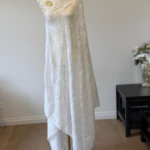 Pu&ograve; includere: Uno scialle o foulard in tessuto bianco semitrasparente drappeggiato su un manichino. Il tessuto presenta un intricato motivo floreale ricamato tono su tono su tutta la superficie, con delicati bordi smerlati lungo l'orlo, creando un aspetto.