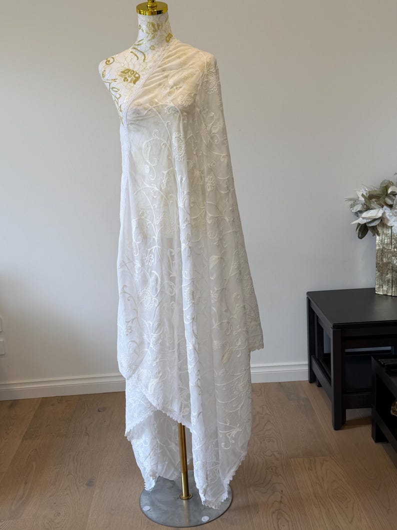 Pu&ograve; includere: Uno scialle o foulard in tessuto bianco semitrasparente drappeggiato su un manichino. Il tessuto presenta un intricato motivo floreale ricamato tono su tono su tutta la superficie, con delicati bordi smerlati lungo l'orlo, creando un aspetto.