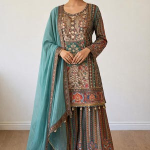 Op de afbeelding: Een ingewikkeld ontworpen teal en bruin salwar kameez ensemble. De outfit heeft gedetailleerde bloemenborduursels, een bijpassende dupatta en een rok met patroon. De top met lange mouwen en de rok hebben een mix van kleuren, waaronder rood, groen en goud.