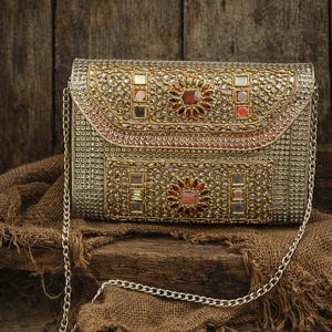 May include: A rectangular, silver-toned clutch bag with detailed decoration. The bag is covered in small, sparkling stones and mirrored squares, with red and gold floral designs. It has a silver chain strap.
