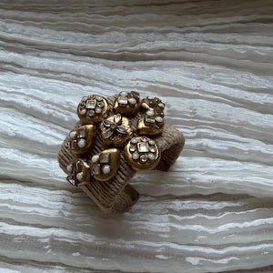 Op de afbeelding: Een goudkleurige manchetarmband versierd met diverse decoratieve knopen. De knopen hebben parel- en kristalaccenten en zijn bevestigd aan een geweven, bruinachtige band. De armband wordt gepresenteerd op een wit, getextureerd oppervlak.