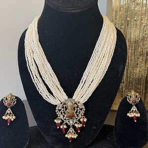 Puede incluir: Un conjunto de collar y pendientes ornamentados, exhibido sobre un busto negro. El collar presenta múltiples hilos de pequeñas cuentas blanquecinas, un colgante detallado con una figura central y detalles rojos y blancos. Los pendientes reflejan el diseño del colgante.