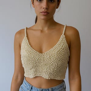 May include: Cream-colored beaded crop top with a V-neckline and spaghetti straps. The top is made of small, round beads, creating a textured surface. The model is wearing light blue denim jeans.