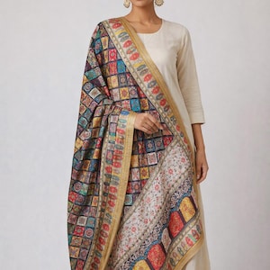 May include: A colorful, patterned shawl with a gold border. The shawl features a geometric design with squares of various colors, including blue, red, yellow, and green. The border has a floral pattern. The shawl is draped over a person wearing a cream-colored outfit.