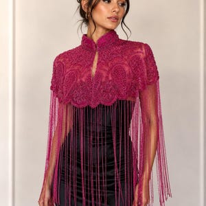 May include: A magenta beaded cape with a high collar and fringe detailing. The cape features intricate beadwork and a flowing fringe that extends down. The garment is worn over a black dress, creating a striking contrast.