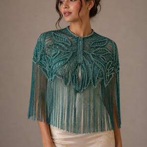 Op de afbeelding: Een teal kralen capelet met een bladeren ontwerp en franjes. De capelet heeft een hoge, ronde halslijn en is versierd met kleine, witte kralen. De franjes hangen naar beneden vanaf de onderkant van de capelet. De capelet wordt gedragen over een crèmekleurige jurk.