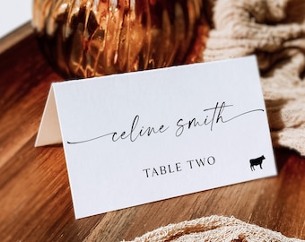 May include: White table card with black text that reads "celine smith" and "TABLE TWO". A small black cow icon is to the right of the text.