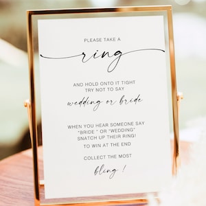 May include: A gold-framed sign with white paper and black text that reads "PLEASE TAKE A ring AND HOLD ONTO IT TIGHT TRY NOT TO SAY wedding or bride WHEN YOU HEAR SOMEONE SAY "BRIDE" OR "WEDDING" SNATCH UP THEIR RING! TO WIN AT THE END COLLECT THE MOST bling!"