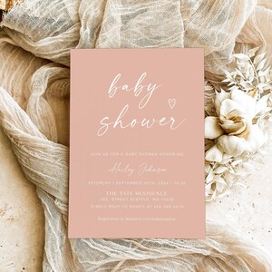 May include: A pink baby shower invitation with white text. The invitation reads "baby shower" with a small heart symbol. The invitation also includes the name "Hailey Johnson", the date and time of the shower, the location, and RSVP information.