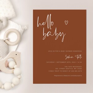 May include: A brown baby shower invitation with white text. The invitation says "hello baby" with a heart symbol. It also includes the name "Sophia Tate", the date and time of the shower, the location, and RSVP information.