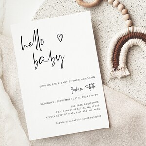 May include: A white baby shower invitation with black calligraphy that reads "hello baby". The invitation includes the details of the event, including the date, time, location, and RSVP information. The text also includes the name "Sophia Tate" and the website "Babylist.com/babysophia".