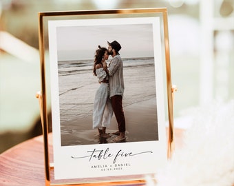 May include: Gold-framed table number sign with a photo of a couple kissing on a beach. The sign reads "Table Five Amelia + Daniel 03.03.2023".