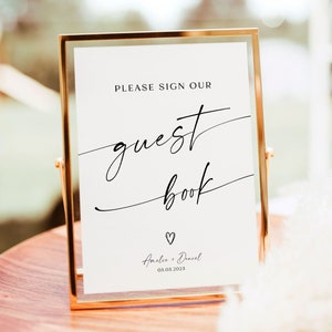 May include: A gold-framed sign with white background and black text that reads "Please sign our guest book" with a small heart and the names "Amelia & Daniel" and the date "03.03.2023".