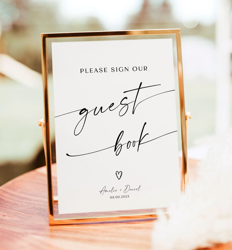 May include: A gold-framed sign with white background and black text that reads "Please sign our guest book" with a small heart and the names "Amelia & Daniel" and the date "03.03.2023".