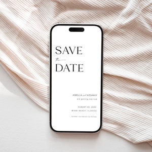 Könnte beinhalten: Ein Smartphone zeigt eine digitale Hochzeitsankündigung. Der Text lautet "SAVE the DATE" in einer minimalistischen Schriftart, mit den Namen Amelia und Kenway, dem Hochzeitsdatum und dem Ort. Das Telefon liegt auf einem neutral getönten Stoff.