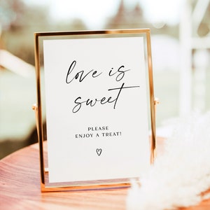 May include: A gold-framed sign with the text "love is sweet" and "Please enjoy a treat!" with a heart symbol. The sign is on a wooden table.