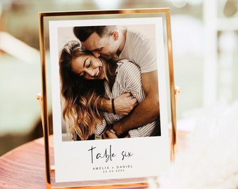 May include: A gold-framed photograph featuring a couple embracing. The photo has a white background with black text that reads "Table Six Amelia + Daniel 03.03.2023".