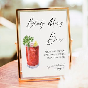 May include: A gold-framed sign with the text "Bloody Mary Bar" in black cursive script. A watercolor illustration of a Bloody Mary cocktail with a celery stalk and lime wedge garnish is next to the text. The sign also includes instructions for making a Bloody Mary: "Pour the vodka, splash some mix, add some kick, + garnish and enjoy!"