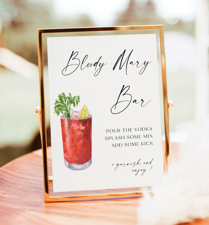 May include: A gold-framed sign with the text "Bloody Mary Bar" in black cursive script. A watercolor illustration of a Bloody Mary cocktail with a celery stalk and lime wedge garnish is next to the text. The sign also includes instructions for making a Bloody Mary: "Pour the vodka, splash some mix, add some kick, + garnish and enjoy!"