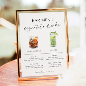May include: A white sign with a gold frame, listing a bar menu with signature drinks. The menu includes an old fashioned, mojito, beer, wine, spirits, and non-alcoholic options. The sign also includes the text "Tara & Kevin 11.09.2022".