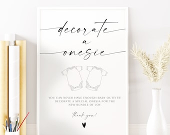 May include: A white sign with black text that says "decorate a onesie" with two outlines of onesies below. The text continues "You can never have enough baby outfits! Decorate a special onesie for the new bundle of joy. Thank you!"