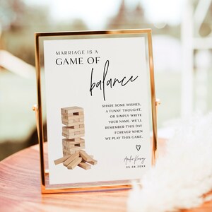 Może przedstawiać: Biały znak z czarnym tekstem, kt&oacute;ry brzmi "Marriage is a game of balance" z rysunkiem gry Jenga i tekstem "Share some wishes, a funny thought, or simply write your name. We'll remember this day forever when we play this game."  Znak znajduje się w złotej ramie i stoi na drewnianym stole. Znak jest podpisany "Aufa-Kry" i datą "25.08.20XX".