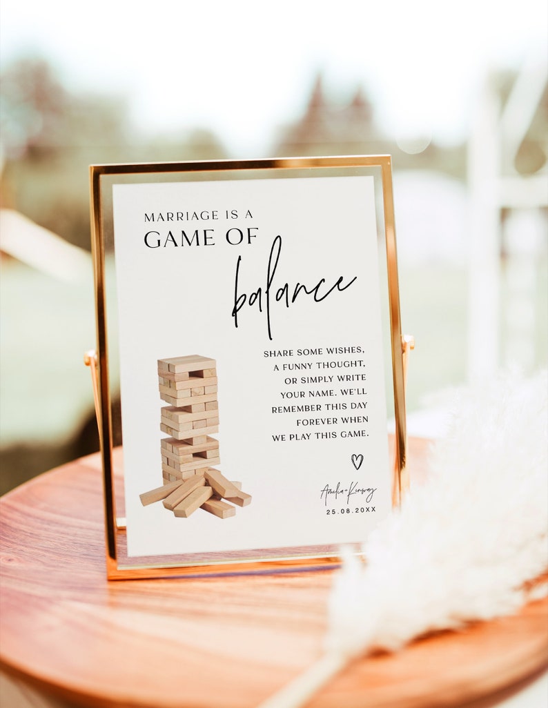 Może przedstawiać: Biały znak z czarnym tekstem, kt&oacute;ry brzmi "Marriage is a game of balance" z rysunkiem gry Jenga i tekstem "Share some wishes, a funny thought, or simply write your name. We'll remember this day forever when we play this game."  Znak znajduje się w złotej ramie i stoi na drewnianym stole. Znak jest podpisany "Aufa-Kry" i datą "25.08.20XX".