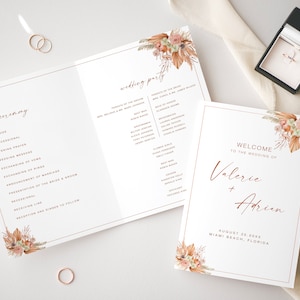 以下が含まれることがあります： 結婚式プログラム。花柄のデザインと「式典」の文字、イベントのリストが記載されています。別途、花柄のデザインと「Valerie + Adrien の結婚式へようこそ」の文字、結婚式の日時と場所が記載されたカードがあります。