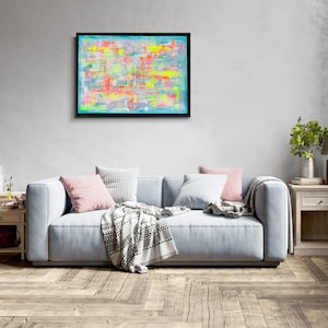 May include: Abstract painting with bright colors, including yellow, pink, and blue, in a black frame hanging on a gray wall above a light blue sofa with pillows and a throw blanket.