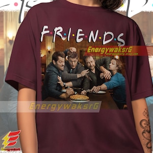 May include: A maroon t-shirt with the words "FRIENDS" in a colorful font, featuring a scene of four men sitting at a table with a pie. The men are all wearing casual clothing and have serious expressions on their faces.