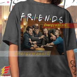 May include: A dark gray t-shirt with the word "FRIENDS" printed in colorful letters. The shirt features a graphic of four men sitting at a table with a pie in front of them. The graphic is in a vintage style.