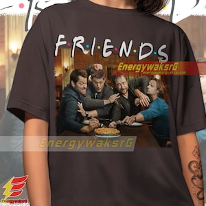 May include: A dark gray t-shirt with the word "FRIENDS" printed in colorful letters. The shirt features a graphic of four men sitting at a table with a pie in the center. The men are all wearing casual clothing and have serious expressions on their faces.