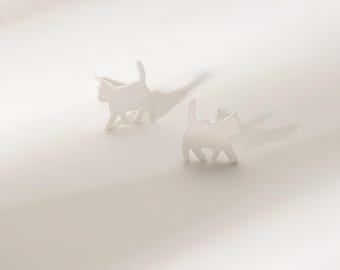 May include: A pair of silver-colored cat-shaped stud earrings. Each earring depicts a walking cat with its tail raised. The earrings are set against a white background, with shadows adding depth.