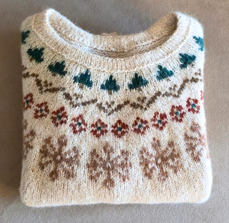 May include: Cream-colored sweater with a round neckline and intricate knit patterns. The design features teal, brown, and red floral and geometric motifs. The folded sweater is a cozy, warm garment.