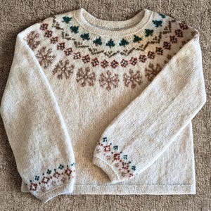 May include: A cream-colored sweater with a crew neck and long sleeves. The sweater features a detailed pattern of brown snowflakes, green trees, and geometric shapes around the yoke and cuffs. The overall design is a classic, cozy style.