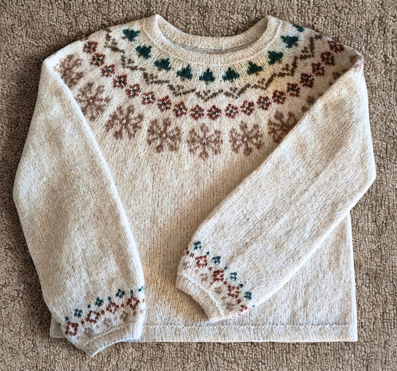 May include: A cream-colored sweater with a crew neck and long sleeves. The sweater features a detailed pattern of brown snowflakes, green trees, and geometric shapes around the yoke and cuffs. The overall design is a classic, cozy style.