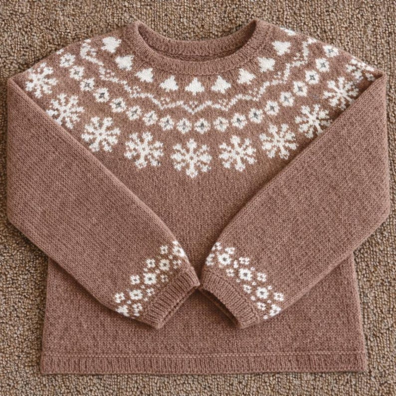 May include: A brown knit sweater with white snowflake and tree patterns across the chest and sleeves. The cuffs also feature a white floral design. The sweater is made of a soft, textured material and is laid flat on a brown surface.