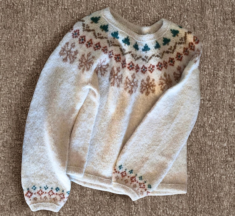 May include: Cream-colored sweater with a crew neck and long sleeves. The sweater features a patterned design in shades of brown, red, and green around the neckline, cuffs, and across the chest. The overall style is a cozy, winter-themed knit.