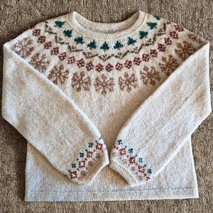 May include: A beige crew neck sweater with a patterned design. The pattern includes brown snowflakes, green trees, and red and brown geometric shapes. The cuffs also have a geometric pattern in green, red, and brown.