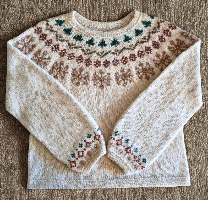 May include: A beige crew neck sweater with a patterned design. The pattern includes brown snowflakes, green trees, and red and brown geometric shapes. The cuffs also have a geometric pattern in green, red, and brown.