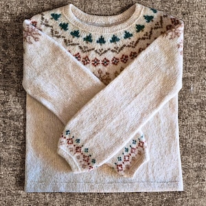 May include: A beige sweater with a crew neck and long sleeves. The yoke and cuffs feature a patterned design in shades of brown, green, and red. The sweater is folded, showcasing the intricate knit details.