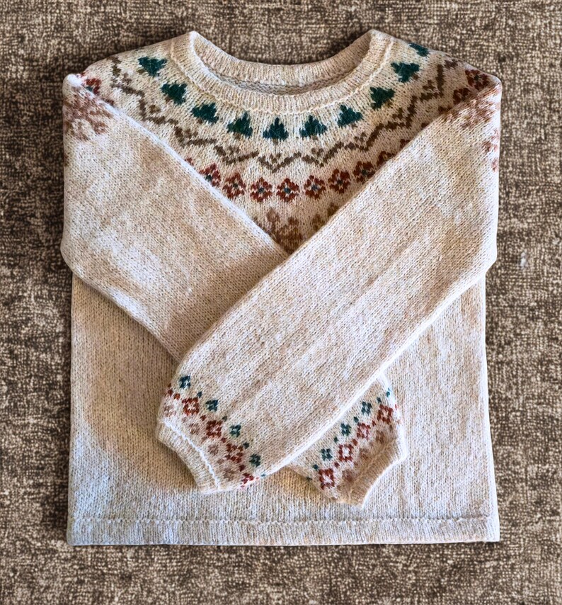 May include: A beige sweater with a crew neck and long sleeves. The yoke and cuffs feature a patterned design in shades of brown, green, and red. The sweater is folded, showcasing the intricate knit details.