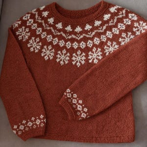 May include: A rust-colored knitted sweater with white snowflake and geometric patterns. The sweater features a crew neck and long sleeves, with the snowflake design across the chest and on the cuffs. The sweater is made of a soft, textured yarn.