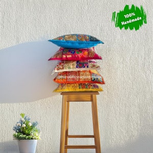 Peut inclure: Une pile de cinq coussins décoratifs faits à la main, colorés, sur un tabouret en bois. Les coussins sont dans les tons de bleu, rose, blanc, orange et jaune, avec des motifs patchwork. Le texte "100% Handmade" est inclus.