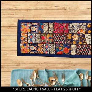 Puede incluir: Un camino de mesa rectangular de patchwork con un borde azul marino. El diseño de patchwork presenta una variedad de patrones florales y geométricos en tonos naranja, rojo, azul y dorado. Cubiertos dorados y una servilleta turquesa en primer plano.