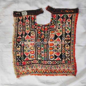 May include: A colorful, embroidered textile with a geometric pattern. The design features red, black, yellow, and blue shapes. The textile has a neckline and is likely a piece of clothing. The text "NY L" is visible.